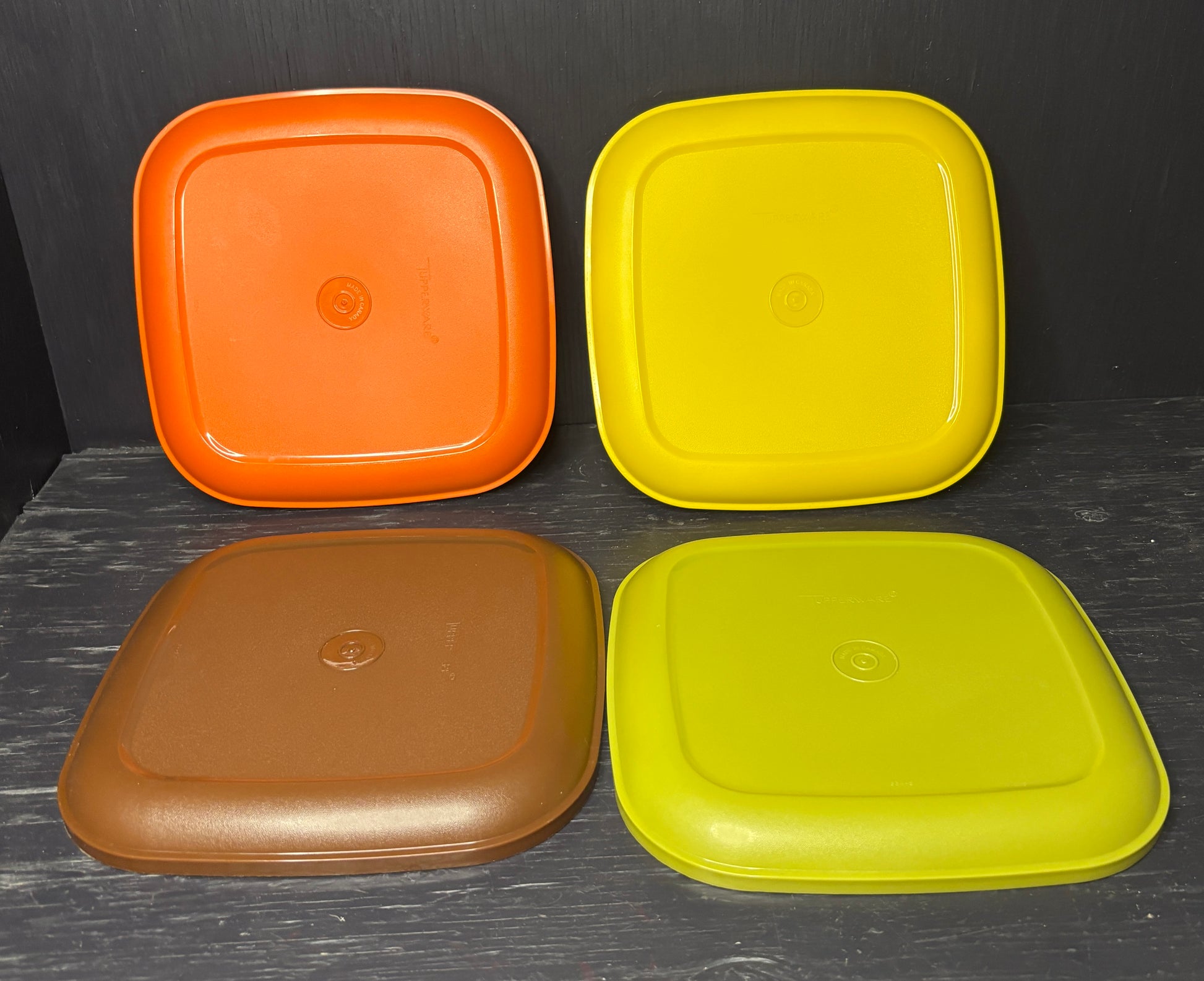 Four square plastic lids in orange, yellow, brown, and green on a dark surface.