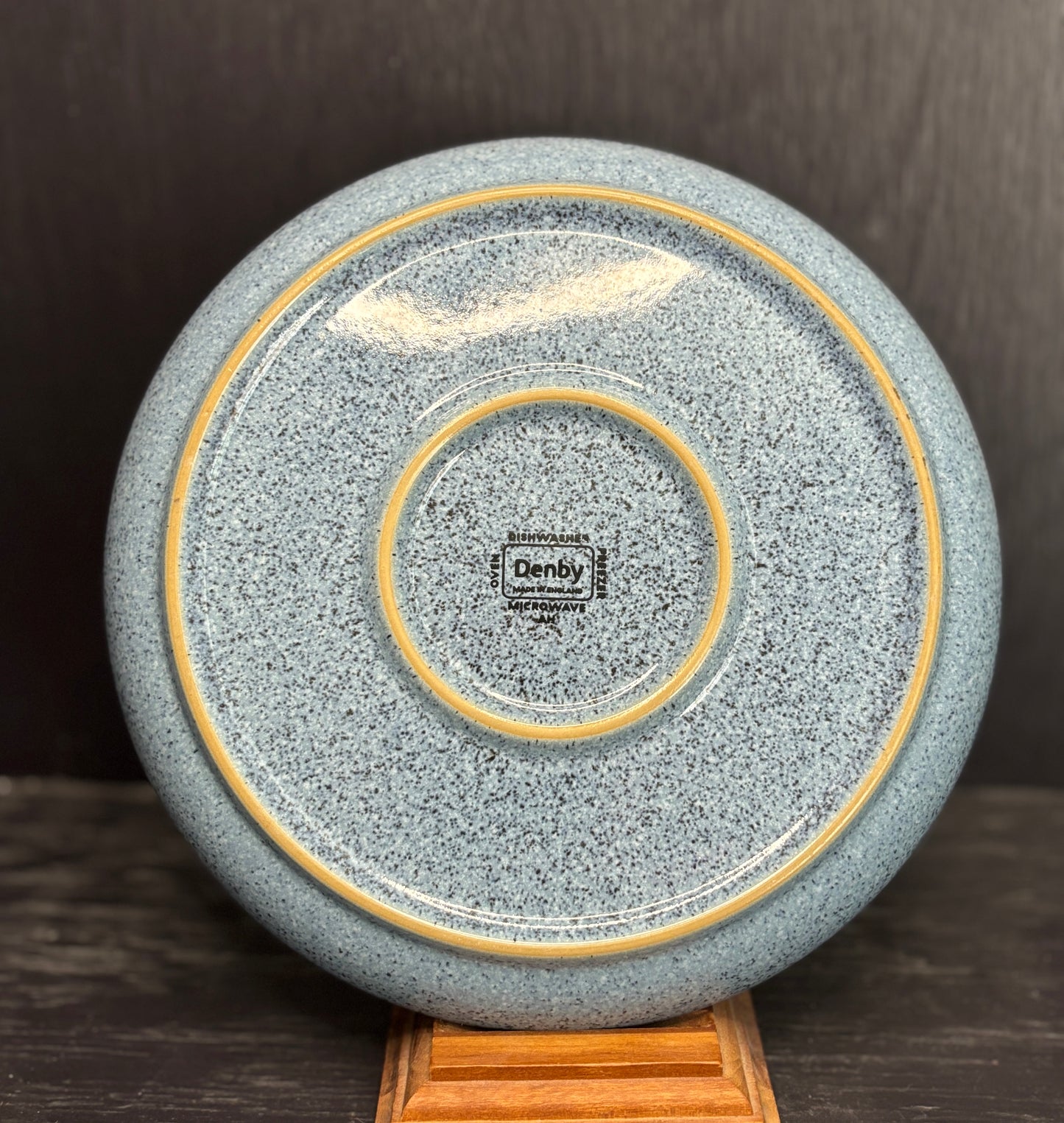 Speckled gray ceramic plate with gold rim on a wooden stand against a dark background