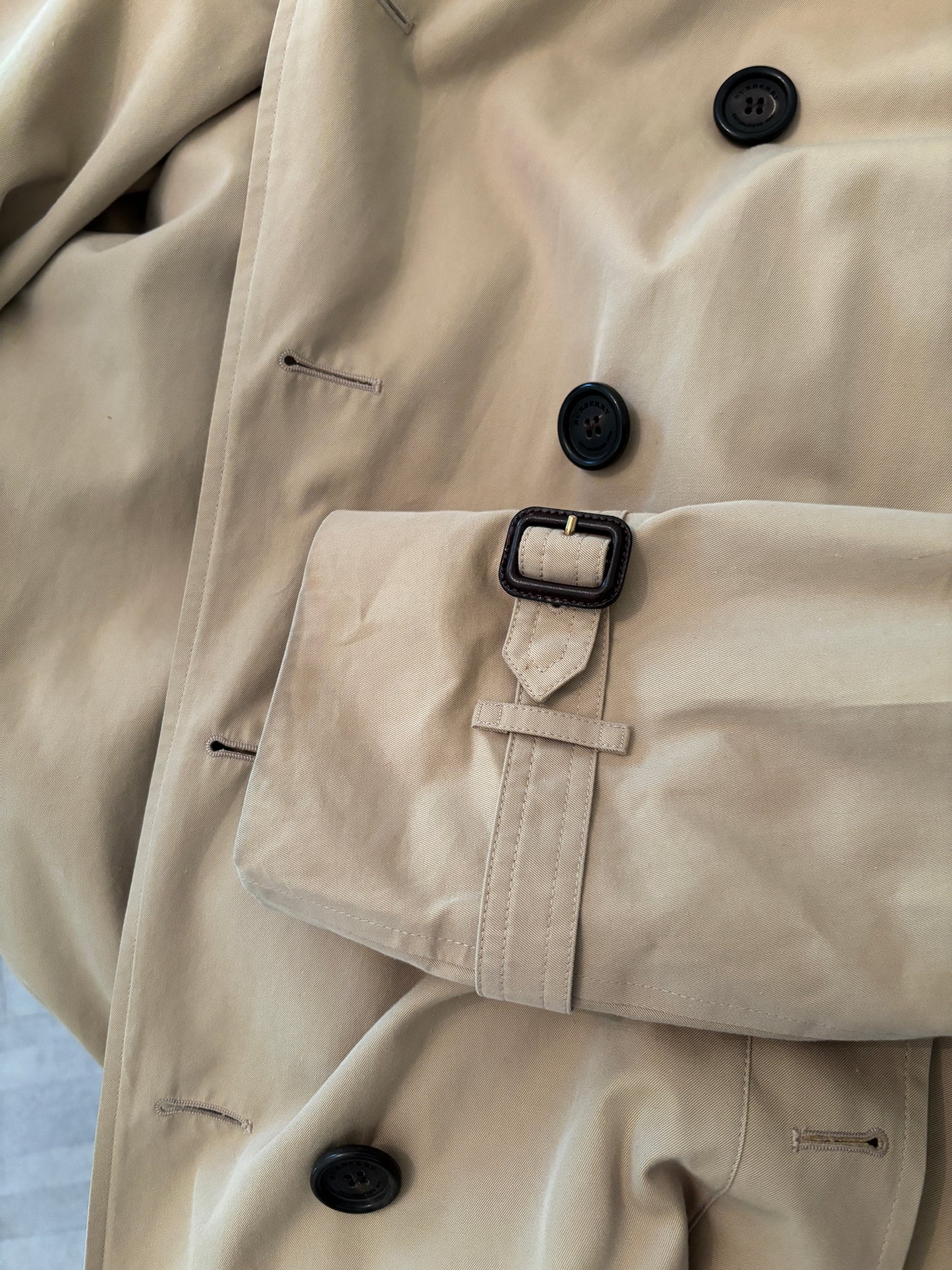 Close-up of a beige trench coat with black buttons and a belt.