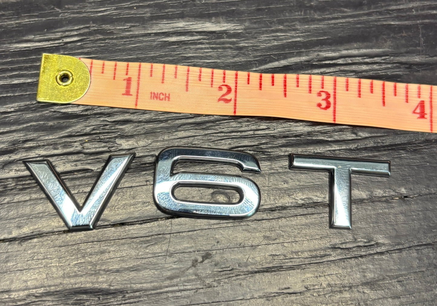 Metallic 'V6T' badge with a measuring tape for scale on a wooden surface
