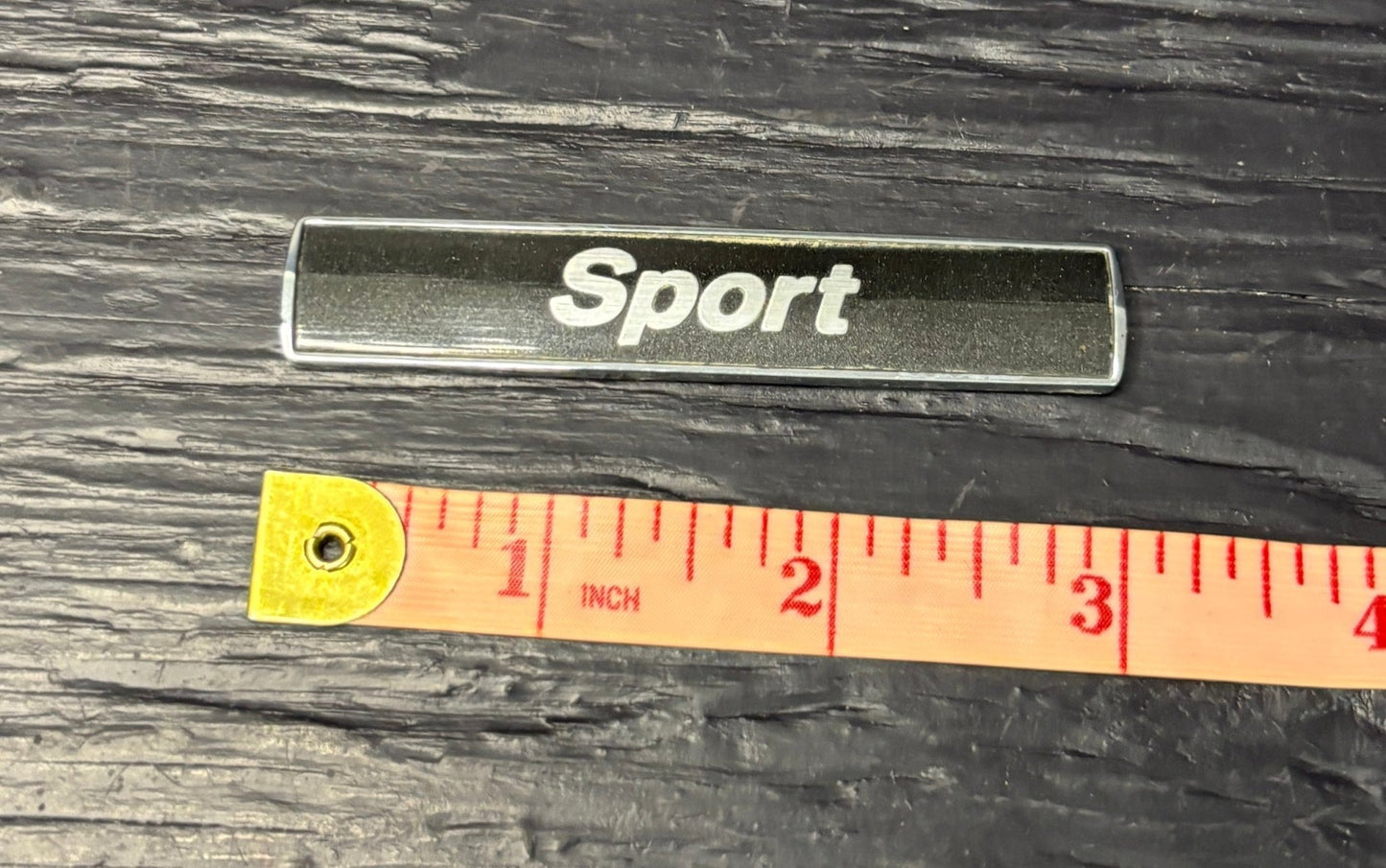 Metallic 'Sport' badge with a ruler for scale on a dark wooden surface