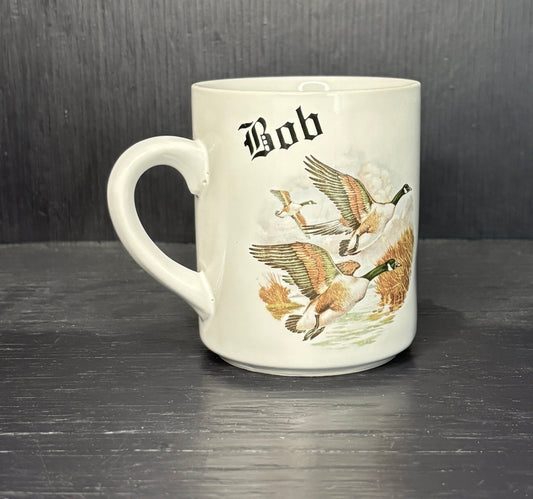 White mug with goose illustrations and 'Bob' text on a dark surface