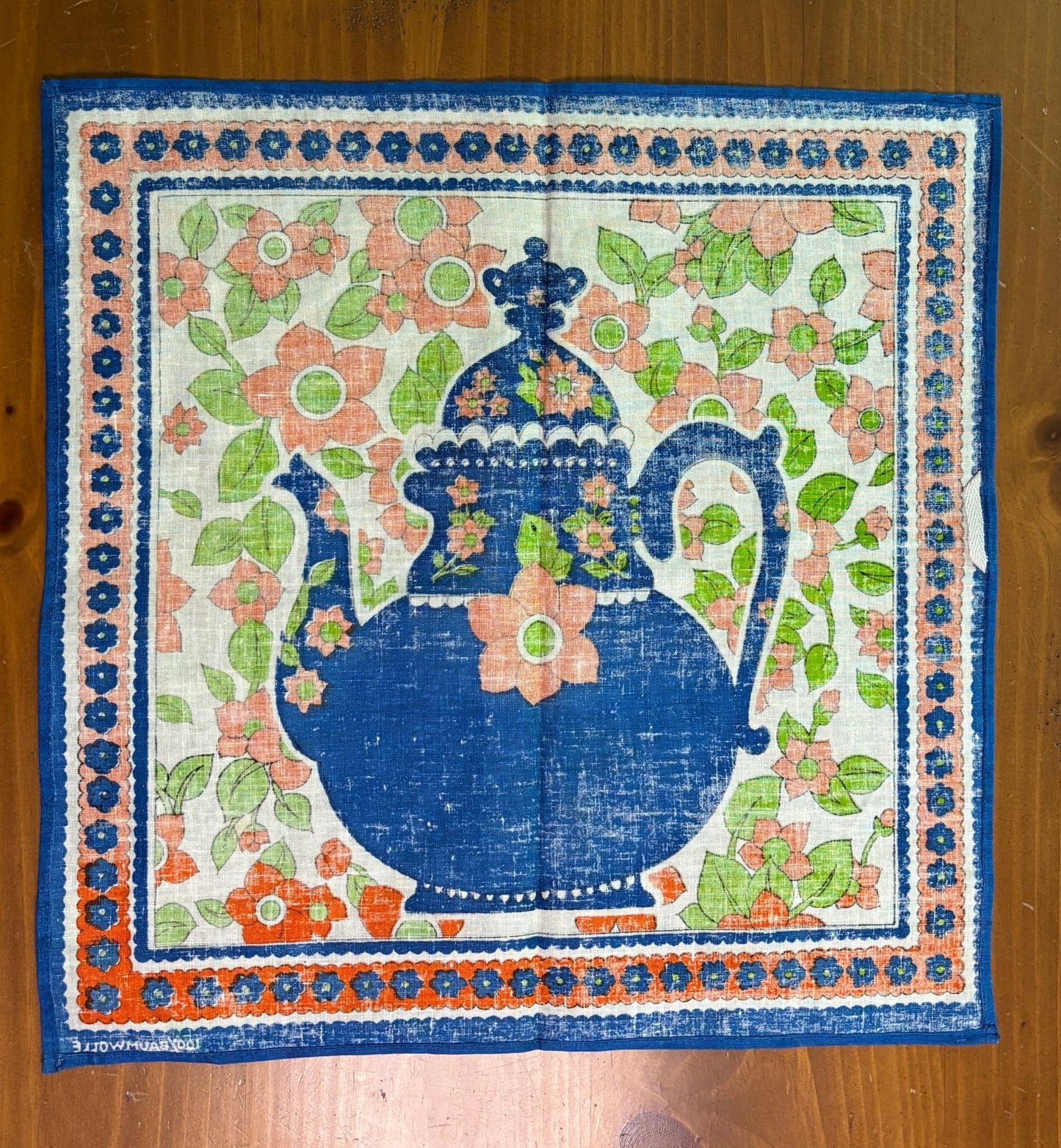 Decorative fabric square with a blue pitcher and floral design on a wooden surface