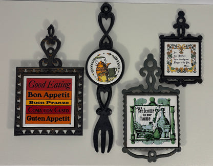 A set of five decorative wall-mounted plates and signs in black metal frames, including food-themed sayings like 'Good Eating Bon Appetit' and 'Welcome to our home', displayed against a light gray wall.