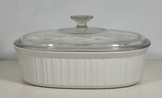 A white ribbed oval ceramic casserole dish with a clear glass lid, photographed against a neutral light gray background.