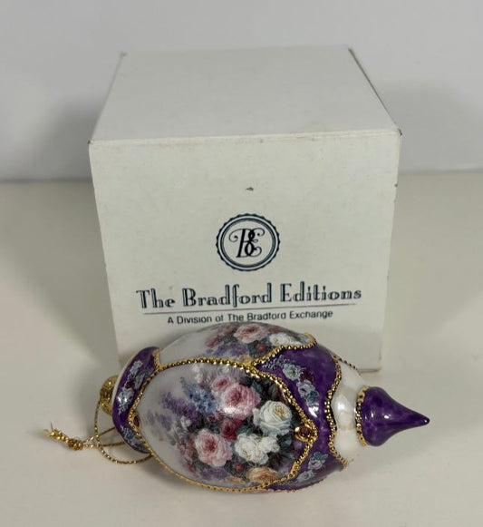 A decorative ceramic ornament shaped like a teardrop or shell, featuring a purple lid, floral painting on a white body, and gold trim, shown in front of its white branded box that reads The Bradford Editions.