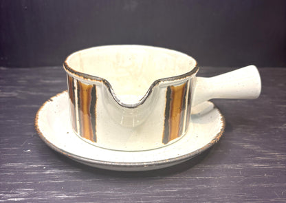 A ceramic gravy boat with a matching saucer, featuring a speckled off-white glaze and vertical metallic gold stripes with dark brown edging, set against a dark background.