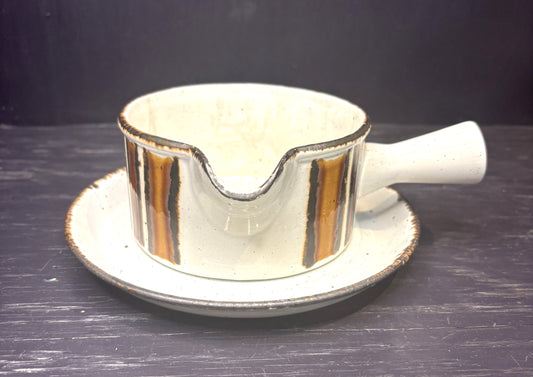 A ceramic gravy boat with a matching saucer, featuring a speckled off-white glaze and vertical metallic gold stripes with dark brown edging, set against a dark background.