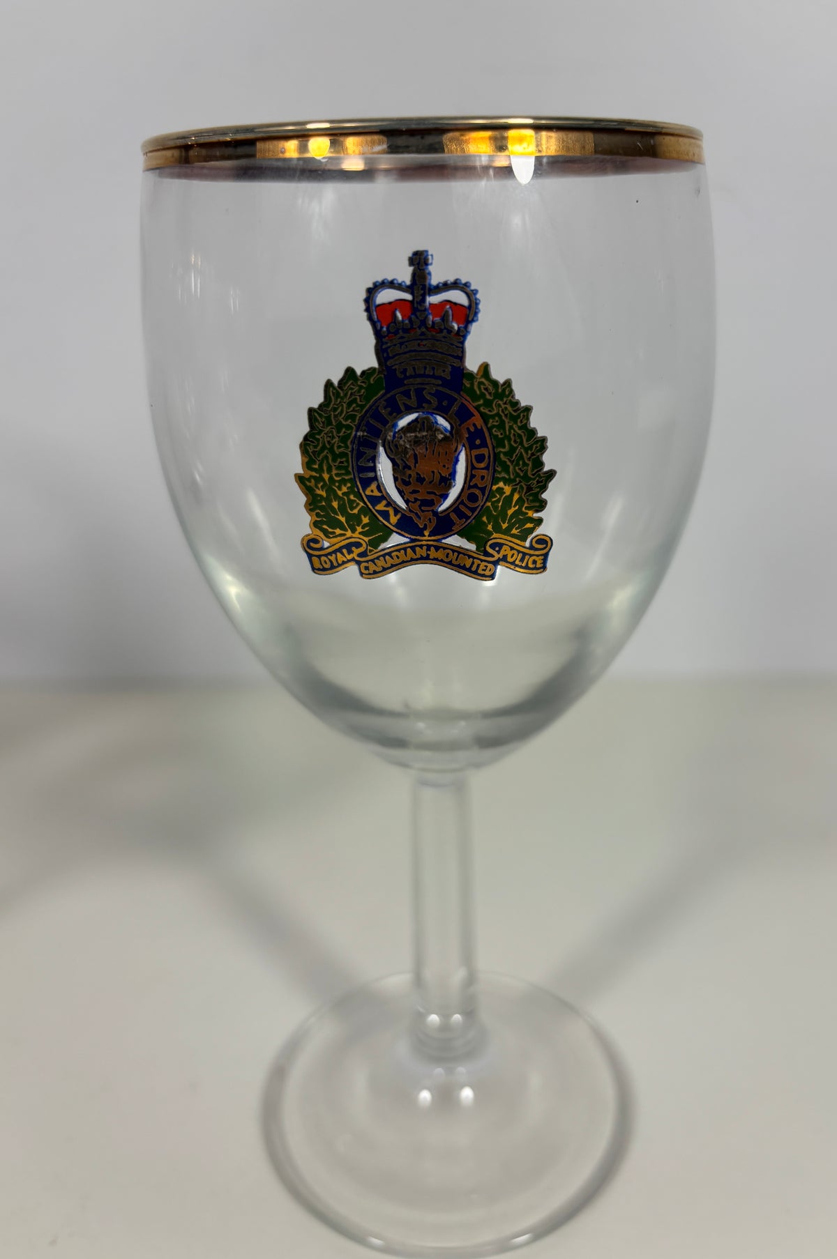 A clear glass stemware goblet with a gold rim and a colorful military-style crest printed on the front, set against a plain light background.