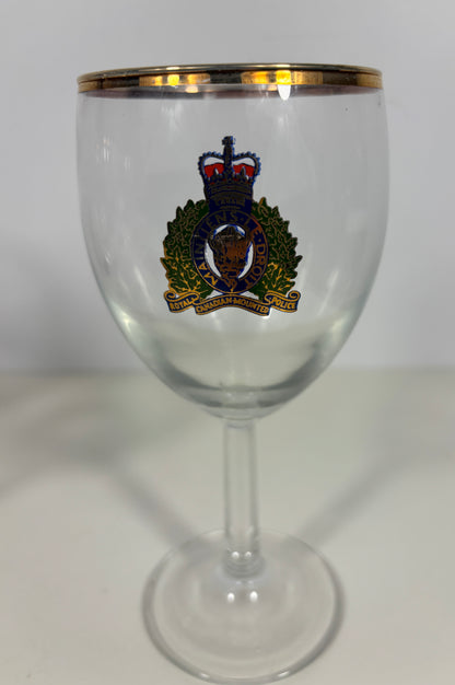 A clear glass stemware goblet with a gold rim and a colorful military-style crest printed on the front, set against a plain light background.