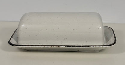 A speckled white ceramic butter dish with a matching lid, featuring a black rim, set against a plain light background.