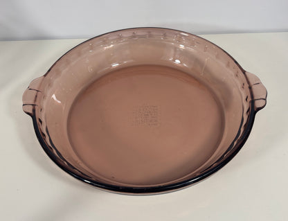 A round, deep glass pie dish in a translucent dusty rose or mauve color, featuring a dark rim and small side handles, photographed on a plain white surface.