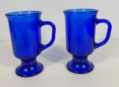 Two vintage cobalt blue glass mugs with pedestal bases and side handles, shown on a plain white background.