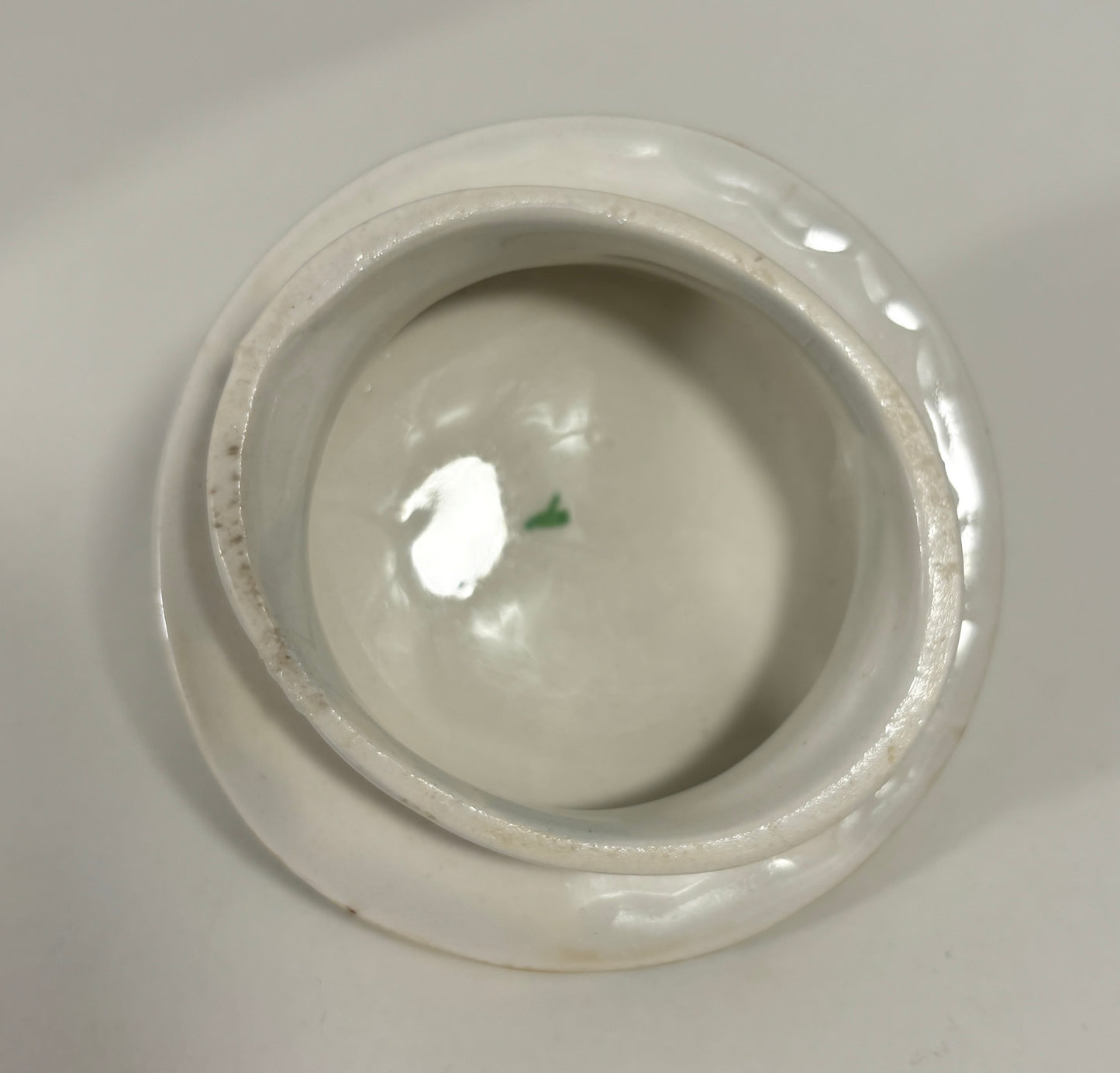 A top-down view of a white ceramic lidded container or small bowl, showing a glossy interior and a slightly worn, rustic rim against a neutral light gray background.