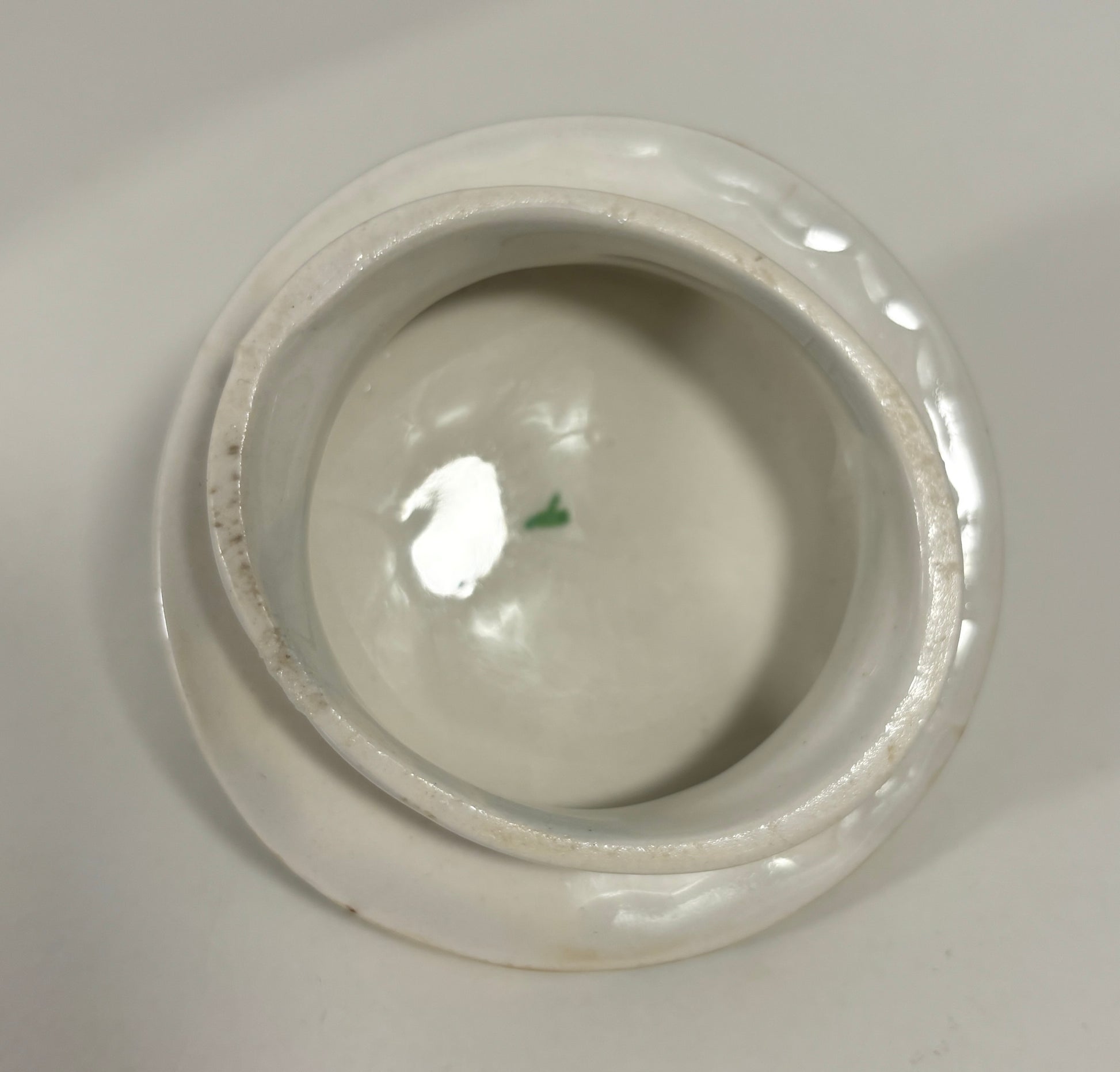 A top-down view of a white ceramic lidded container or small bowl, showing a glossy interior and a slightly worn, rustic rim against a neutral light gray background.