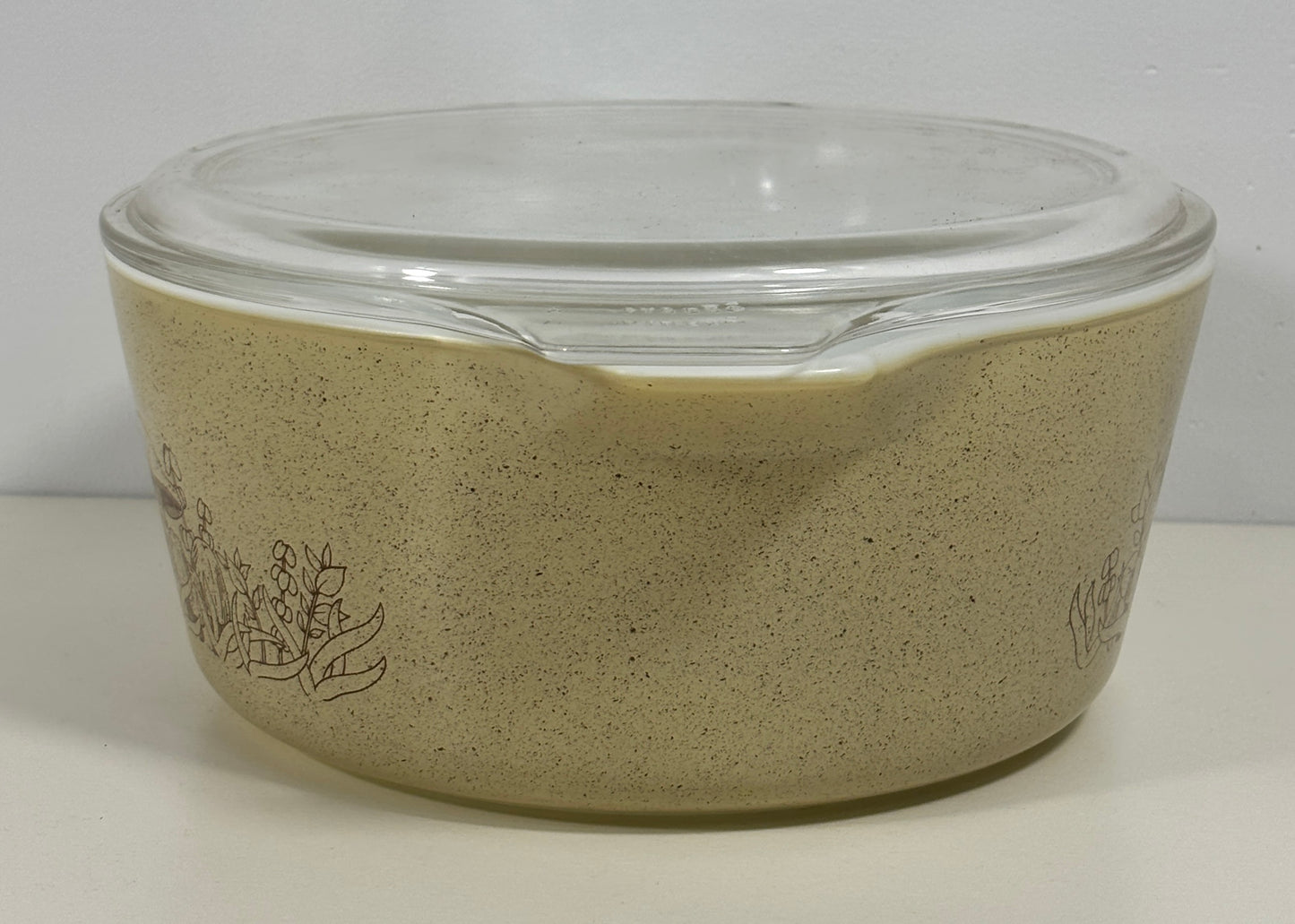 A vintage Pyrex glass casserole dish with a clear glass lid, featuring a speckled beige exterior and a dark brown floral line drawing on the side.
