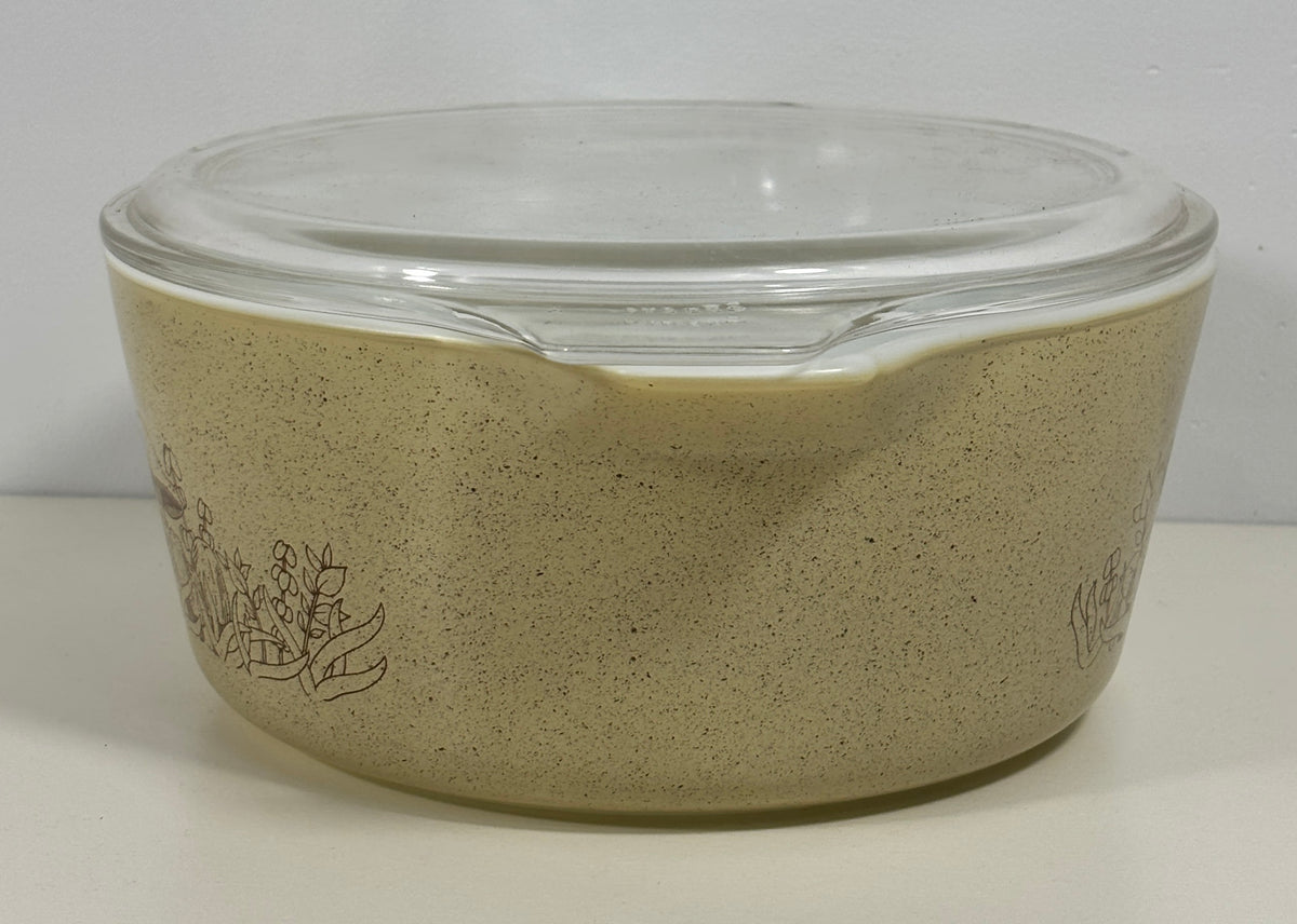 A vintage Pyrex glass casserole dish with a clear glass lid, featuring a speckled beige exterior and a dark brown floral line drawing on the side.