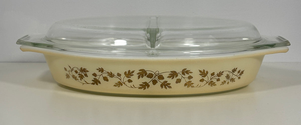 A vintage Pyrex oval casserole dish with a cream-colored body, decorated with a brown floral vine pattern, and topped with a clear glass lid.