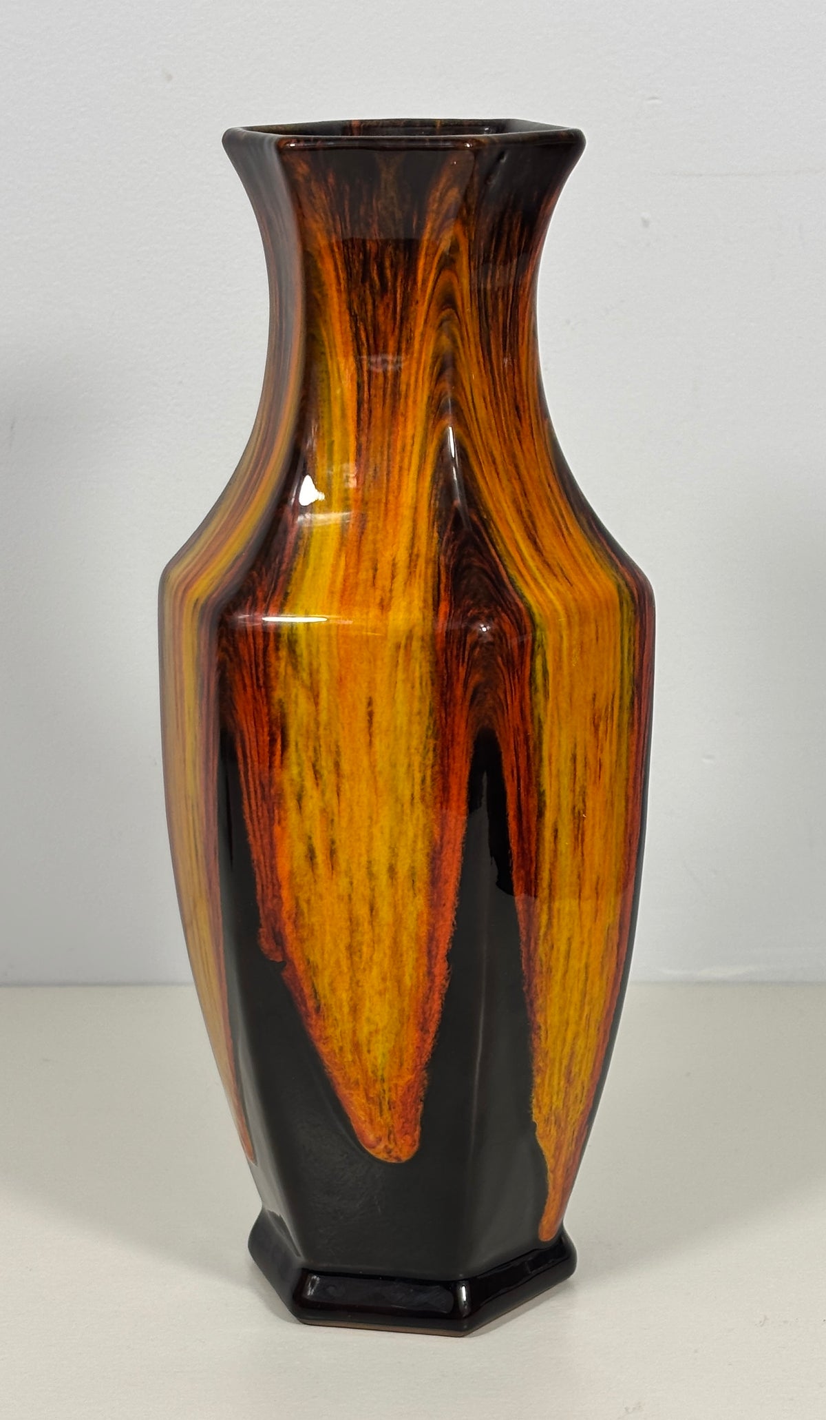 A glossy ceramic vase with a hexagonal base, featuring a marbled wood-grain pattern in vibrant orange, yellow, and deep brown over a black background.