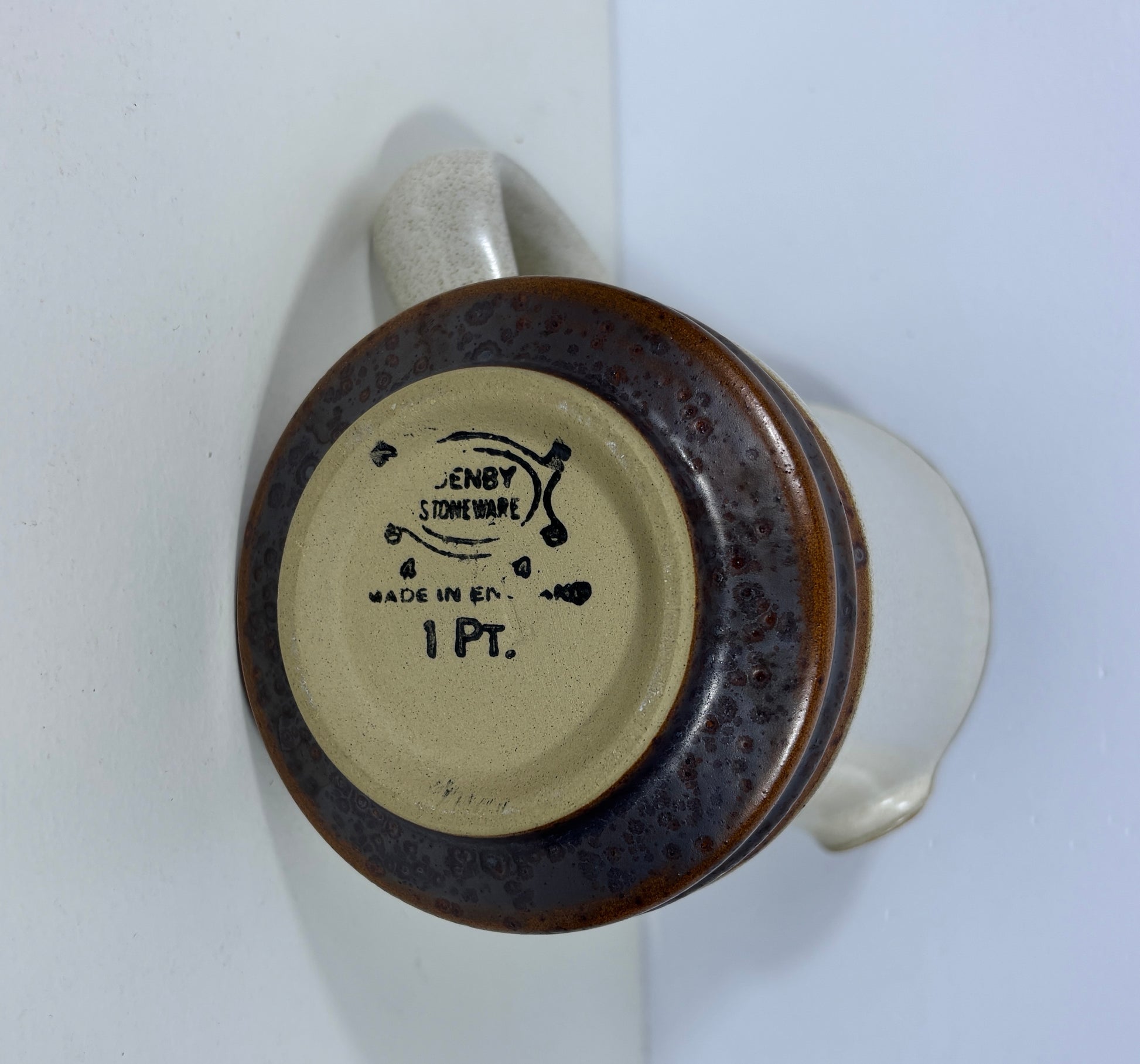 The bottom of a ceramic mug or pitcher showing a dark brown speckled glazed rim, a beige center with the brand 'Denby STONEWARE' and '1 PT.' printed in black, and a light cream-colored handle and body visible on the right.