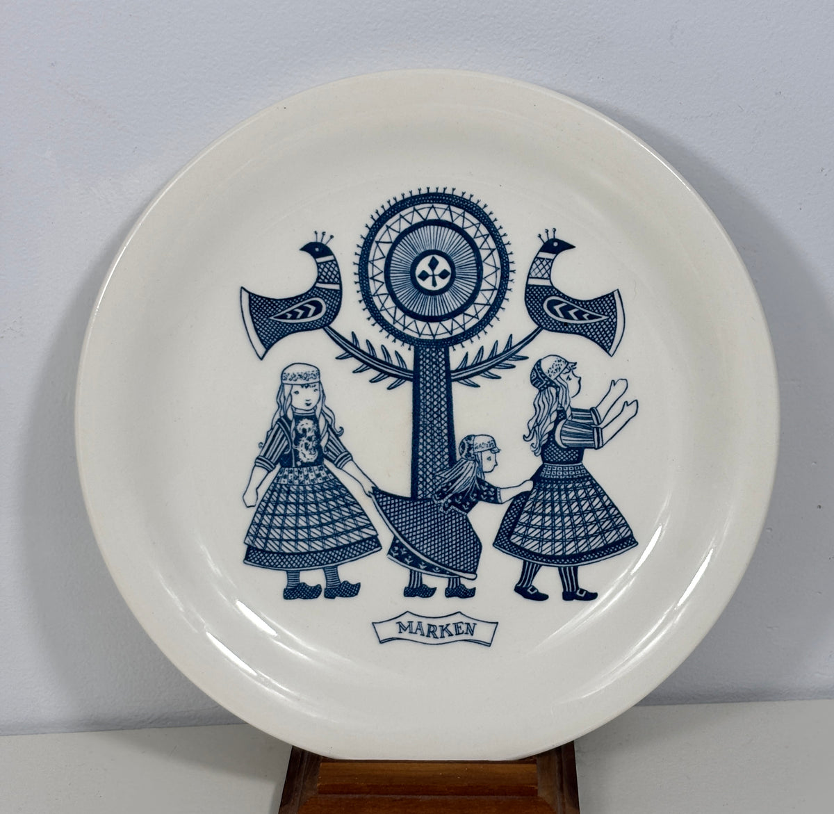 A white ceramic decorative plate featuring a blue printed illustration of three girls, a central tree motif, birds, and a banner reading 'MARKY'. The plate is displayed on a wooden stand against a plain light gray wall.