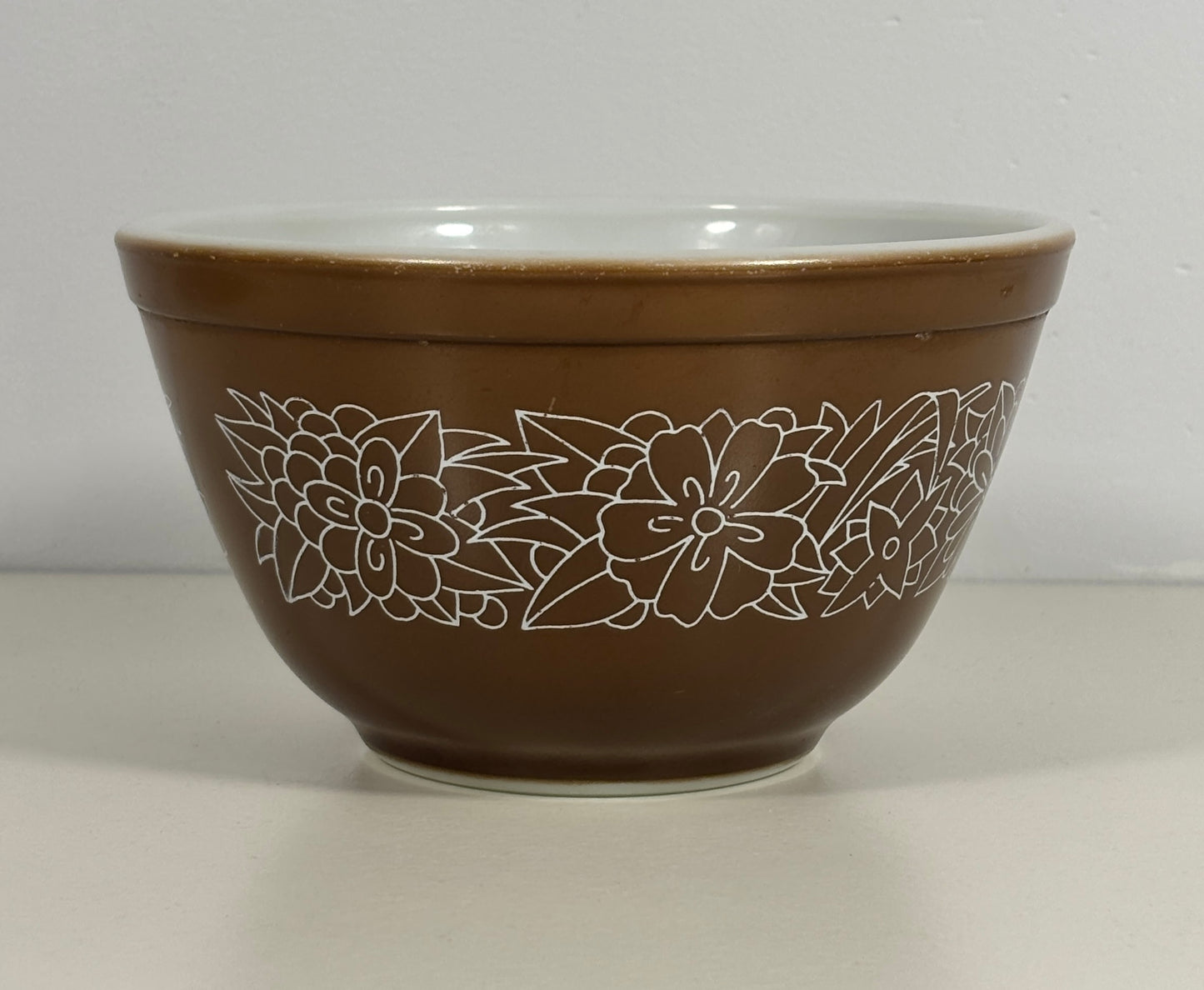 A brown mixing bowl with a white floral line pattern around the outside and a white interior, shown against a neutral light gray background.