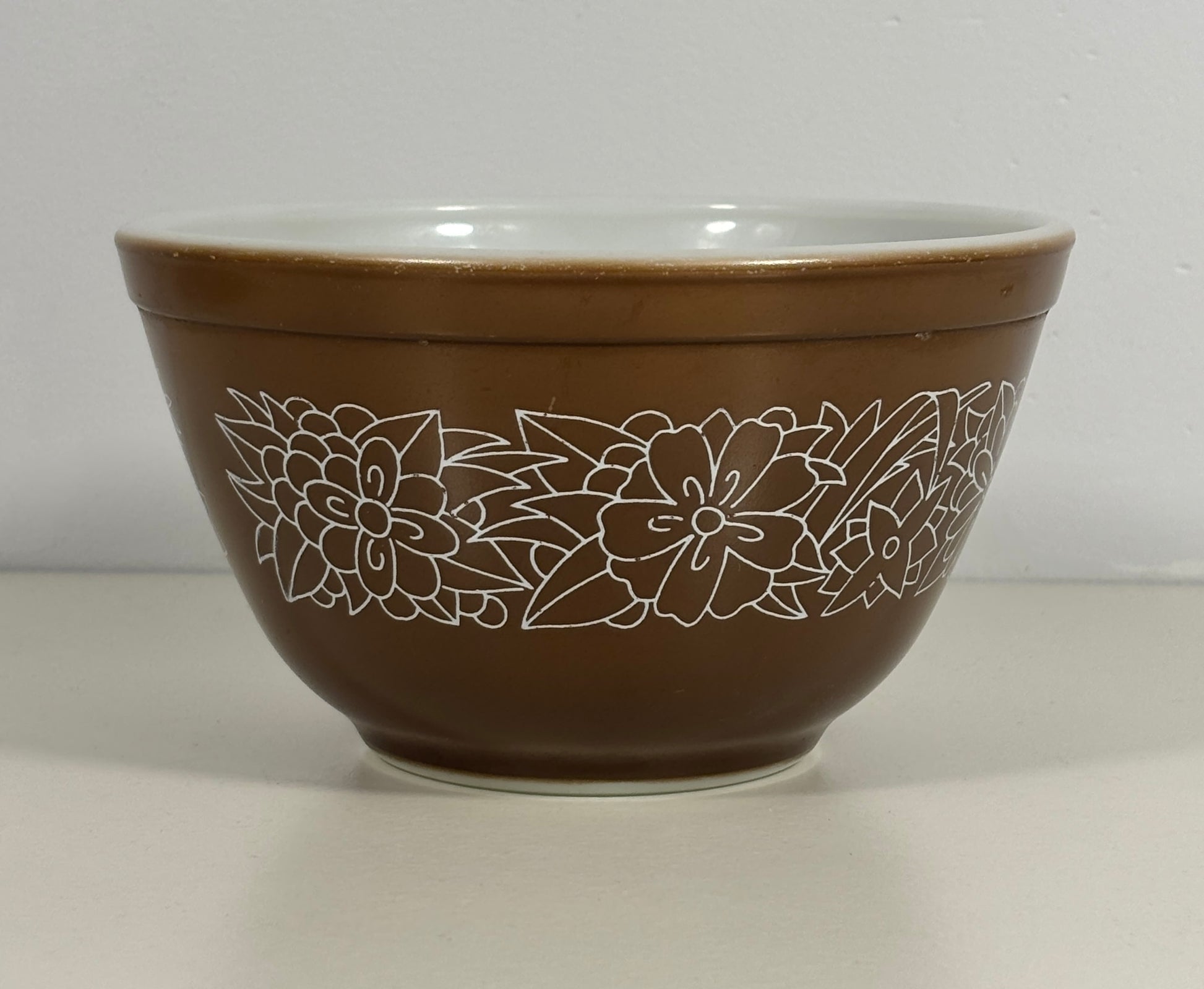 A brown mixing bowl with a white floral line pattern around the outside and a white interior, shown against a neutral light gray background.