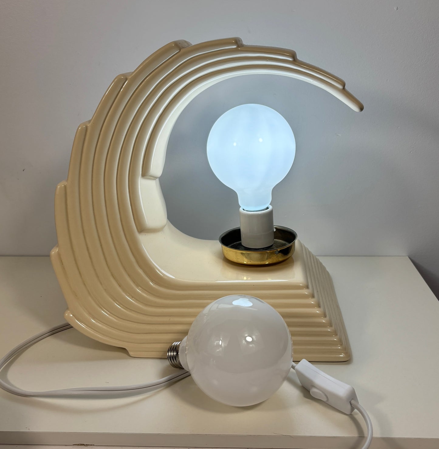 A retro-style wave-shaped table lamp in cream-colored ceramic, shown lit with a glowing bulb inside. A second unlit bulb and its power cord are placed in front on a white surface against a plain light gray wall.
