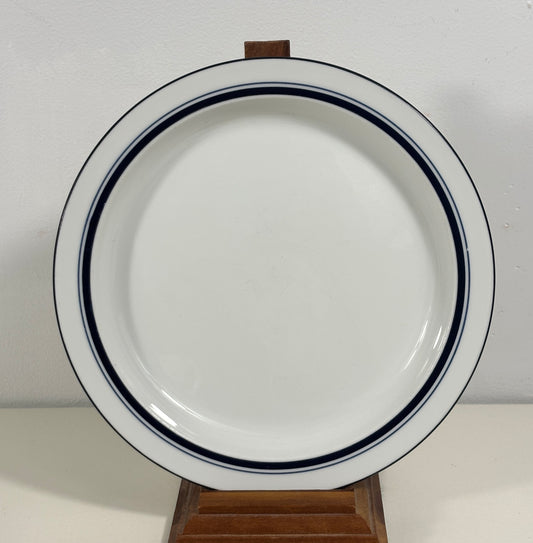 A white ceramic plate with two dark blue stripes along the rim, displayed on a small wooden stand against a plain light background.