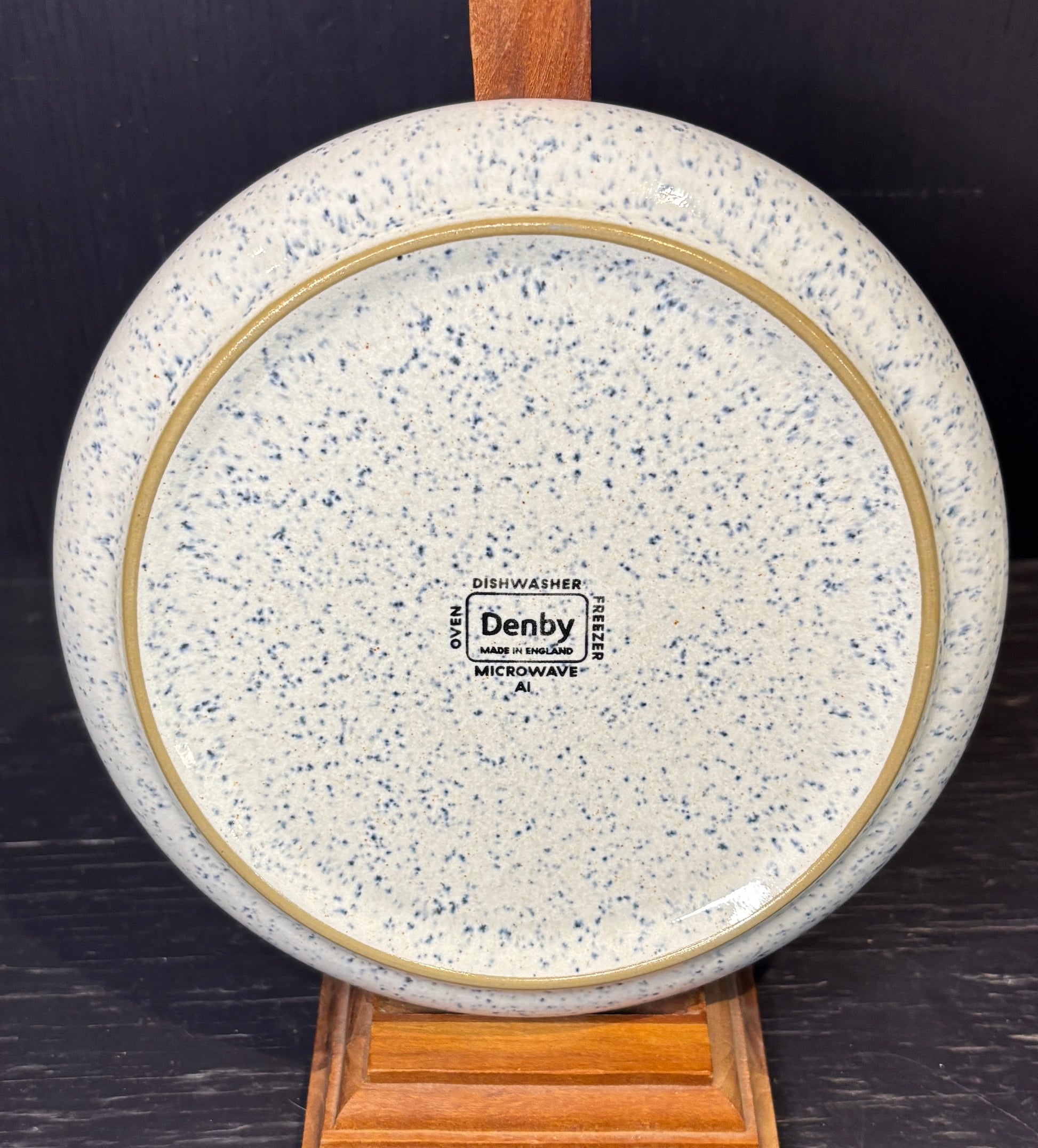 Speckled ceramic plate with 'Denby' branding on a wooden stand against a dark background