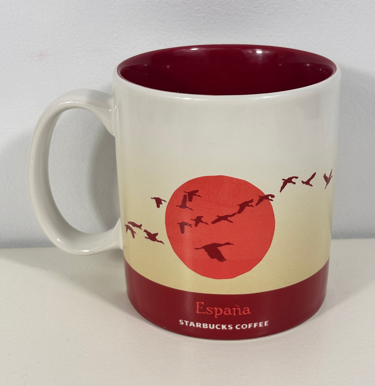 A white ceramic Starbucks Coffee mug with a dark red interior and base, featuring a cream-to-red gradient design with a red sun and flying birds, and the text 'España STARBUCKS COFFEE' on the front.