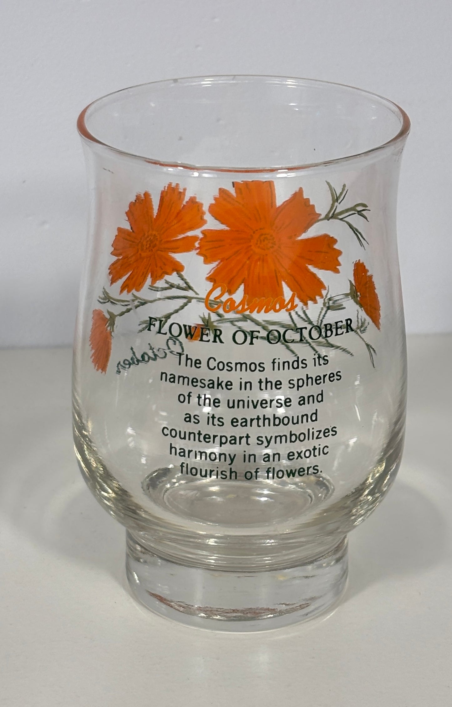 A clear glass tumbler with a thick base, decorated with orange cosmos flowers and black text reading 'Flower of October' and a poem about the cosmos, photographed against a plain white background.