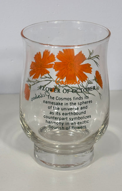 A clear glass tumbler with a thick base, decorated with orange cosmos flowers and black text reading 'Flower of October' and a poem about the cosmos, photographed against a plain white background.