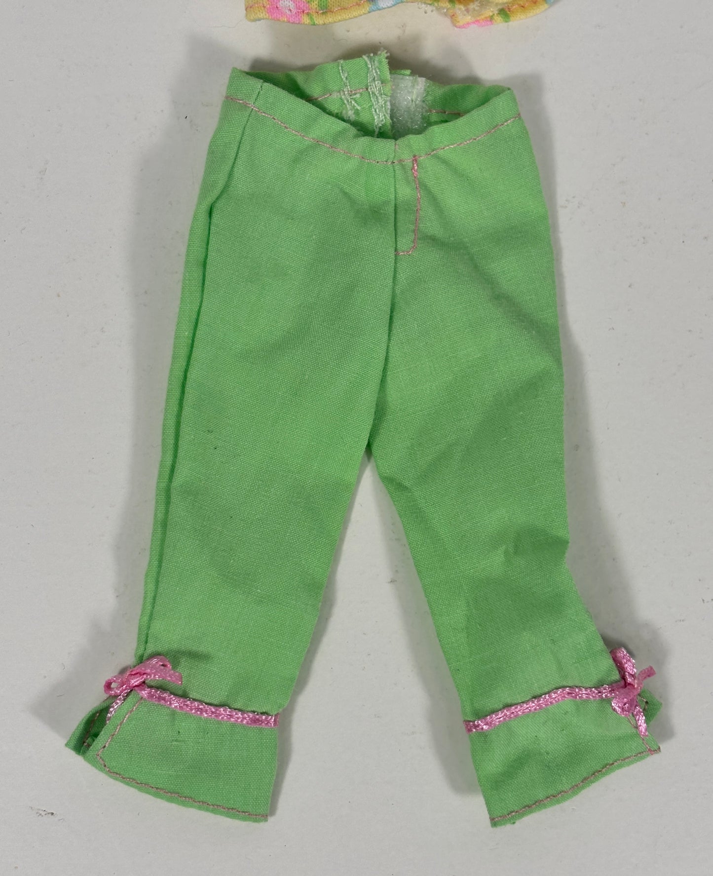 Green pants for dolls, featuring pink glitter trim and small pink bows on the cuffs, laid flat on a light gray background.
