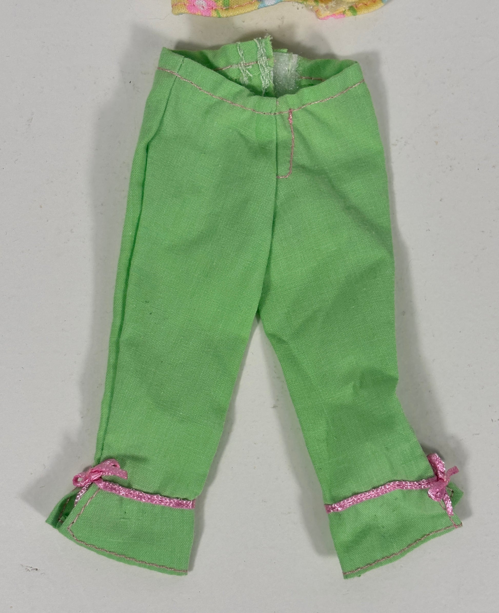 Green pants for dolls, featuring pink glitter trim and small pink bows on the cuffs, laid flat on a light gray background.