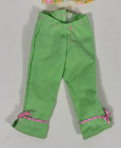 Green pants for dolls, featuring pink glitter trim and small pink bows on the cuffs, laid flat on a light gray background.
