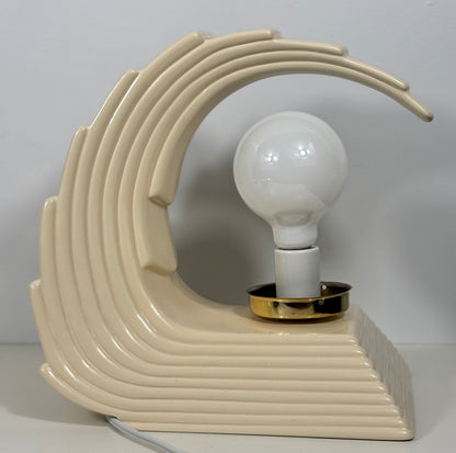 A retro-style ceramic table lamp with a sculpted wave-like form in cream beige, featuring a white globe bulb and a gold-finished socket ring, set against a plain light gray background.