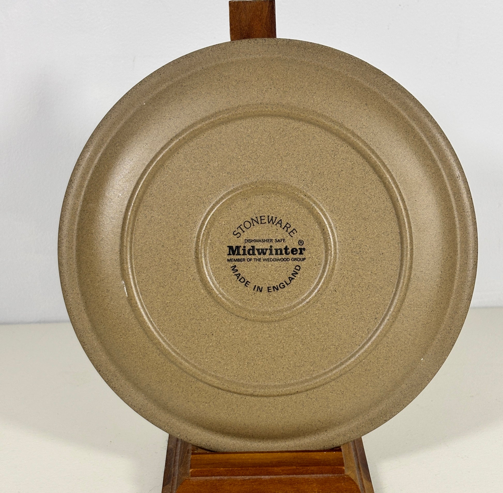A round beige speckled ceramic plate or shallow dish shown from the back, displayed on a wooden stand against a plain white background. The center features a circular printed label that reads 'STONeware midwinter'.