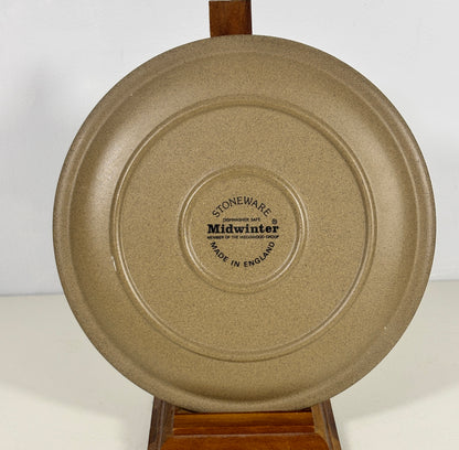 A round beige speckled ceramic plate or shallow dish shown from the back, displayed on a wooden stand against a plain white background. The center features a circular printed label that reads 'STONeware midwinter'.