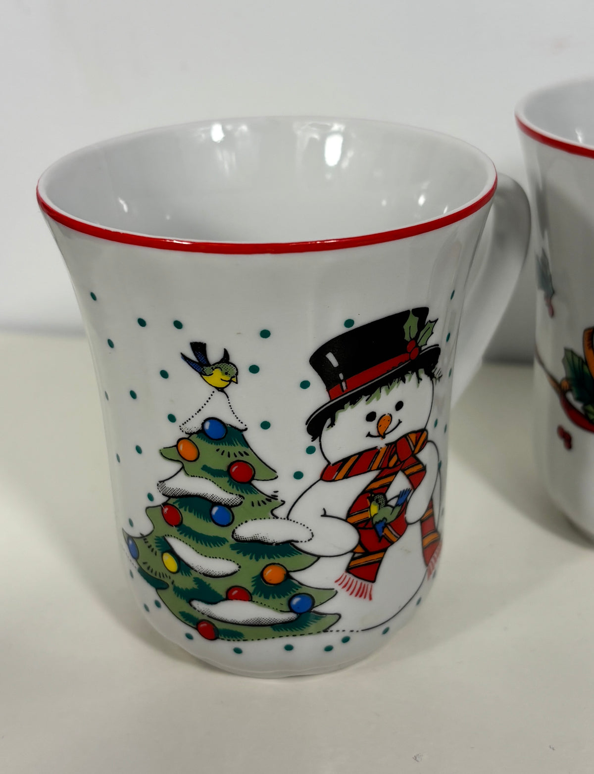 A white ceramic mug with a red rim, decorated with a festive winter scene featuring a snowman in a black top hat and red striped scarf next to a decorated Christmas tree with colorful ornaments.