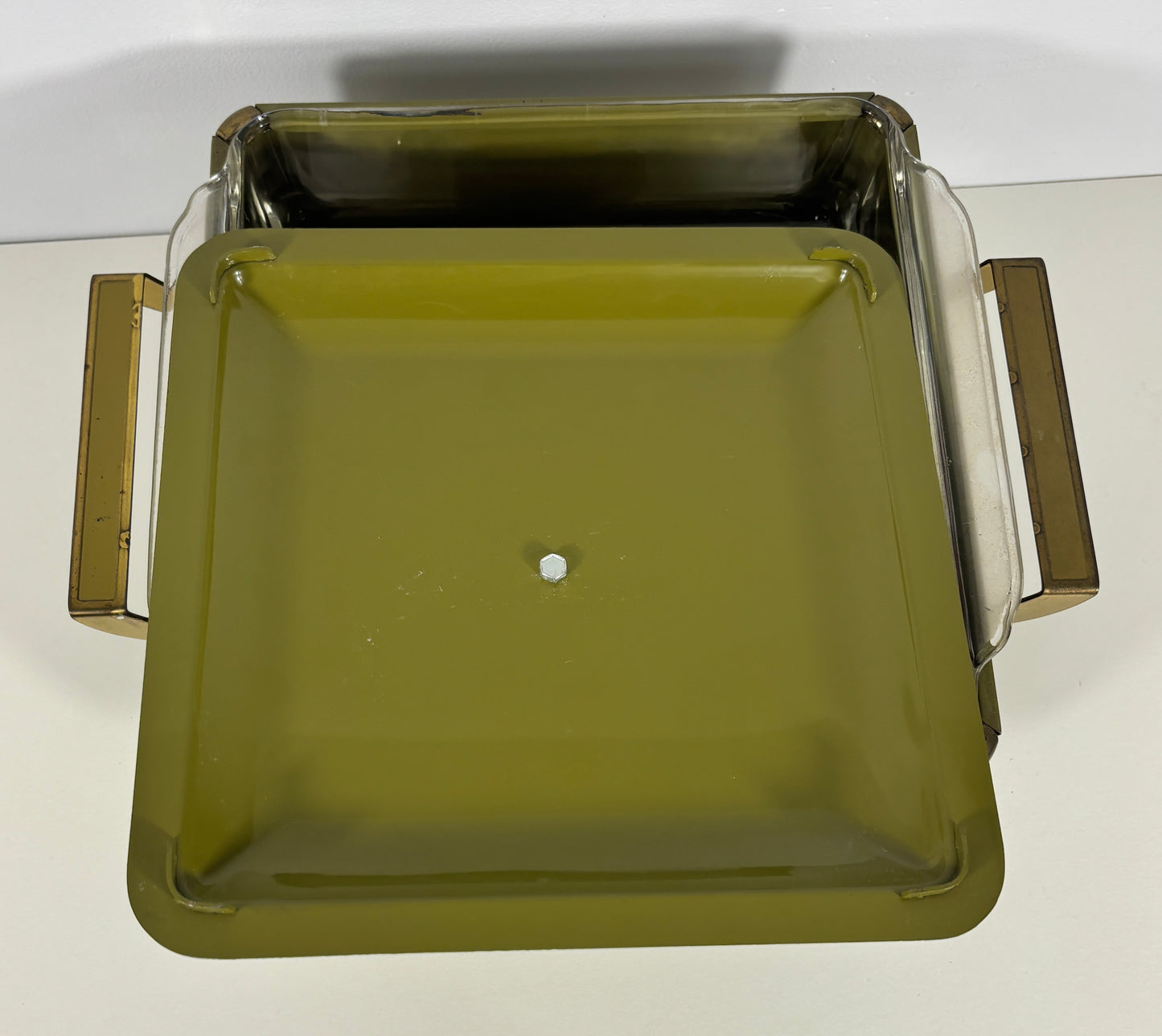 An olive green square glass baking dish with clear glass sides and gold-toned metal handles, photographed from a top-down angle on a white surface.