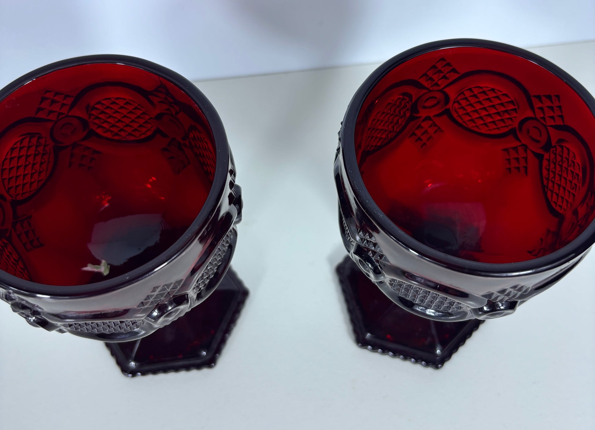A pair of vintage red glass goblets with dark red rims and textured cut glass detailing, displayed on a light surface.