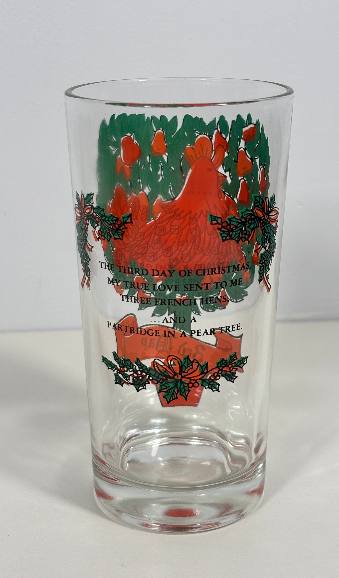 A clear glass pint glass featuring a vintage Christmas design with a red rooster, green holly, and red ornaments, alongside printed text.