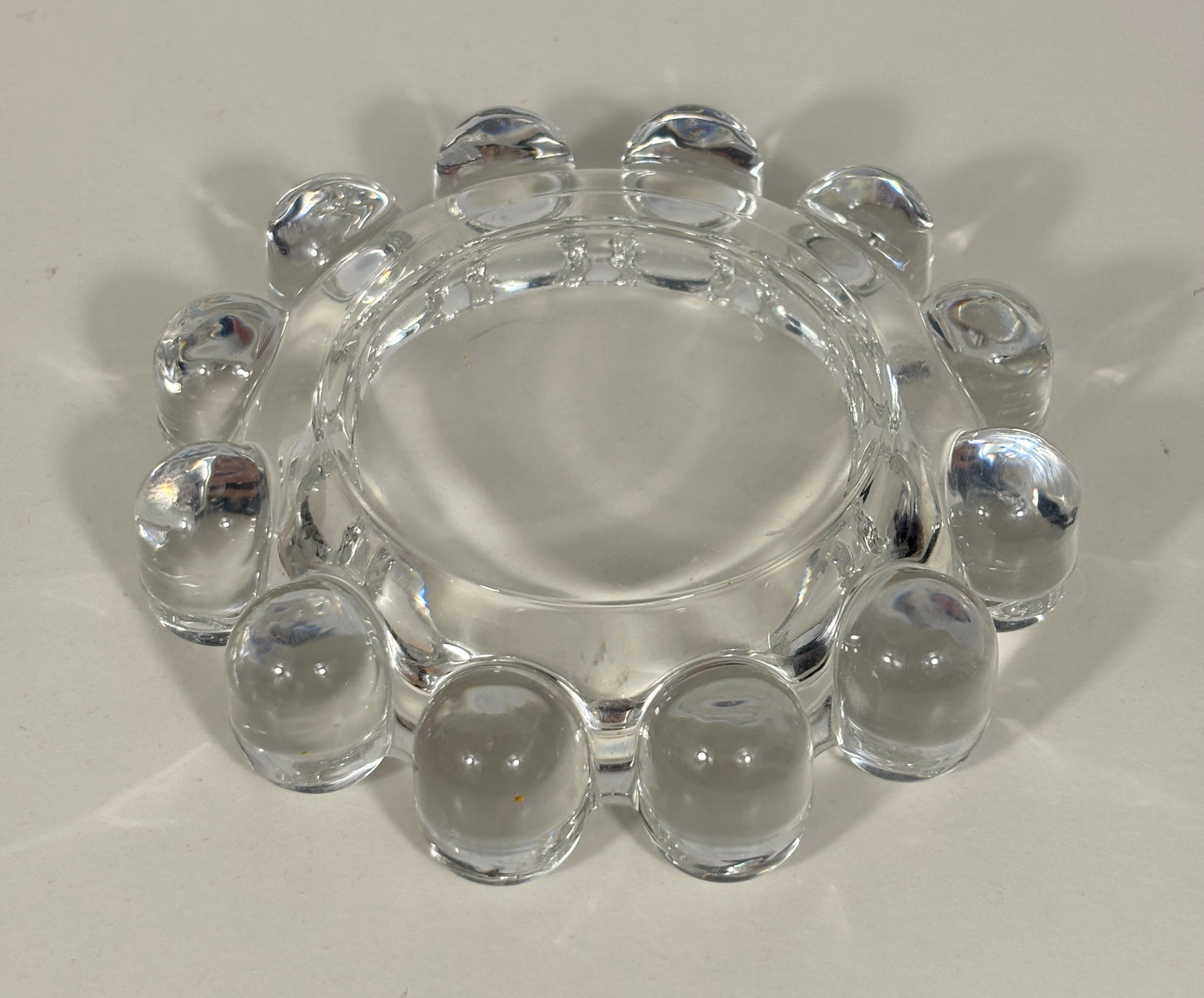 Clear glass ashtray with a circular center and a ring of raised bubble-like grips around the edge, photographed on a light neutral background.