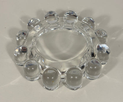 Clear glass ashtray with a circular center and a ring of raised bubble-like grips around the edge, photographed on a light neutral background.