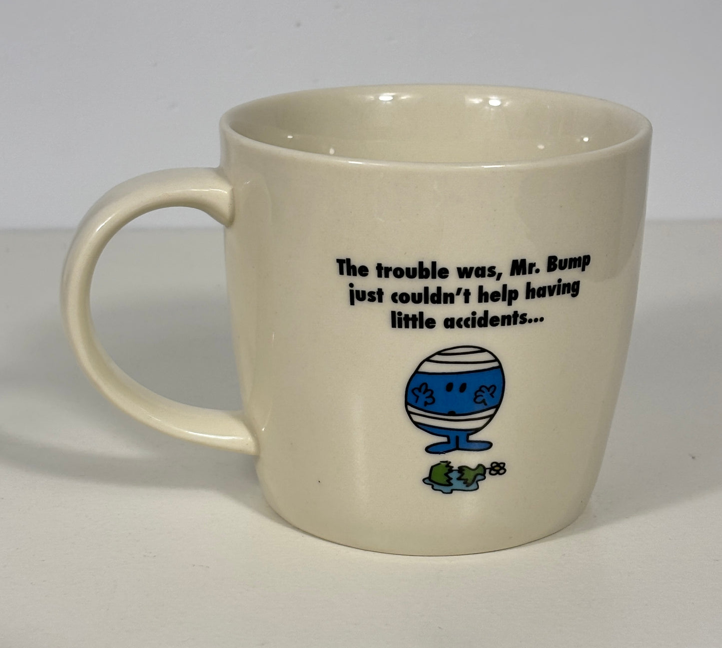 An off-white ceramic mug featuring a black printed quote and a blue-and-white Mr. Bump cartoon illustration with a small green frog below, set against a plain light background.