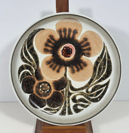 A round ceramic decorative plate with a mid-century modern floral design, displayed on a wooden stand against a plain white wall.
