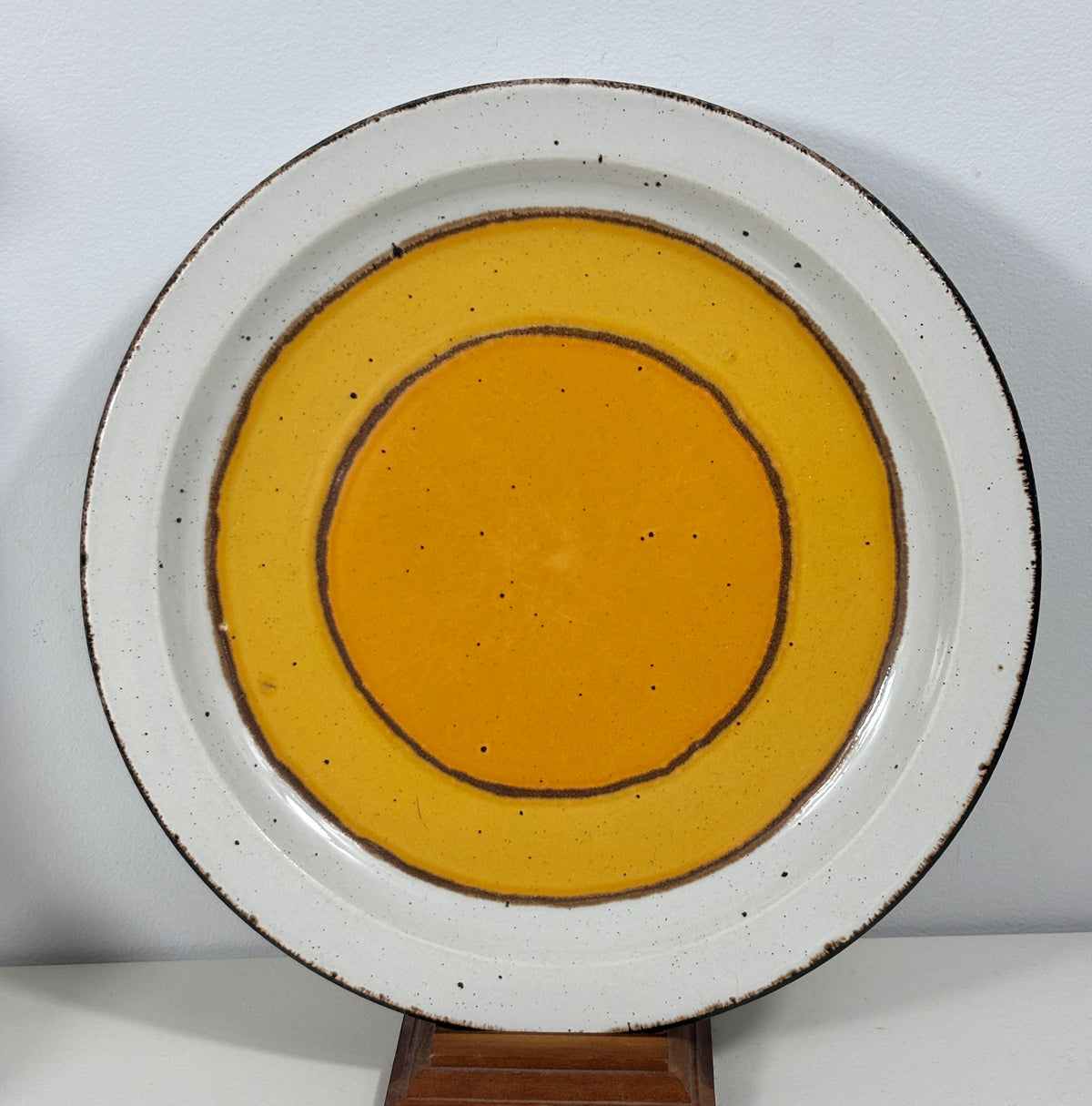 A round ceramic plate with a white speckled rim and concentric yellow-orange glazed circles, displayed on a small wooden stand against a plain light background.