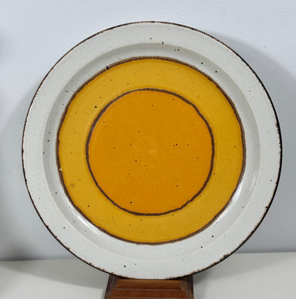 A round ceramic plate with a white speckled rim and concentric yellow-orange glazed circles, displayed on a small wooden stand against a plain light background.