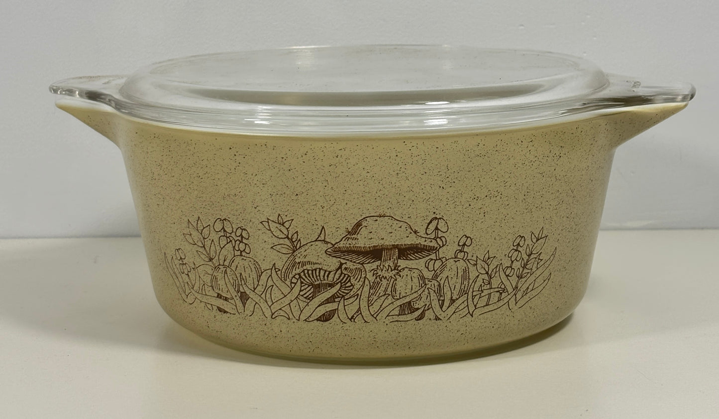 A vintage Pyrex casserole dish with a clear glass lid, featuring a speckled beige body with a brown mushroom and wildflower pattern, set against a plain light background.